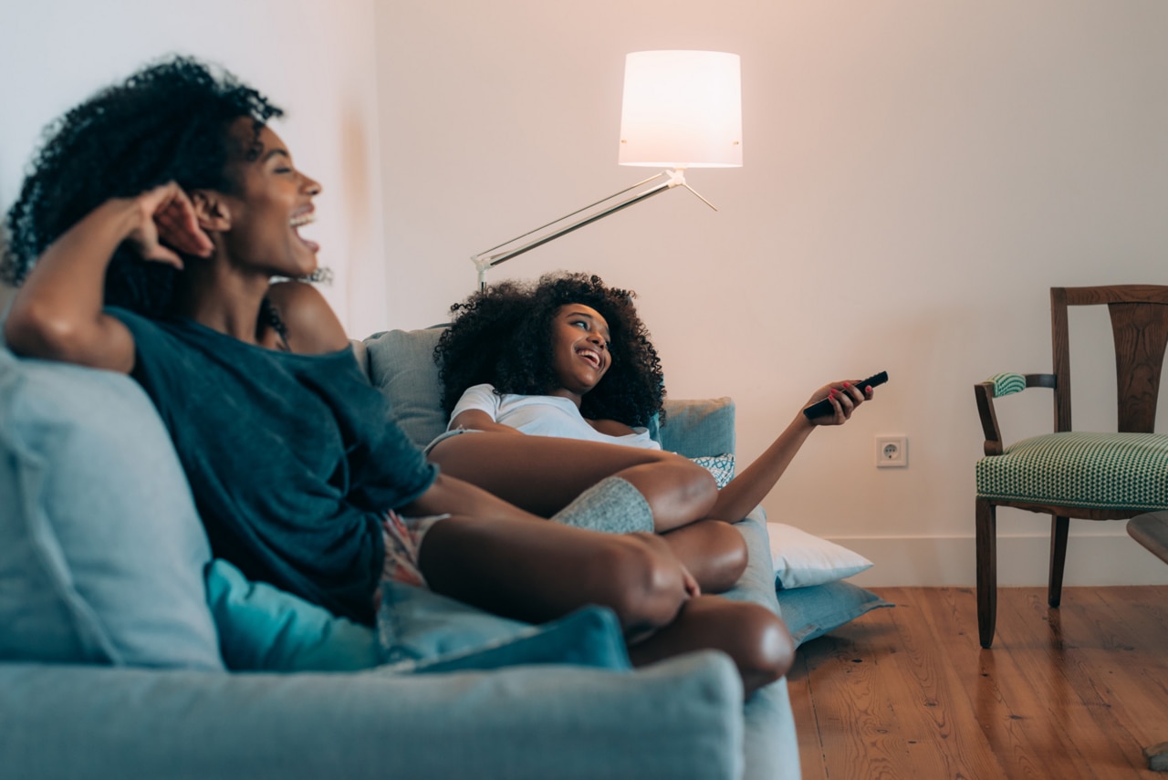 Happy young two black women lying down in the couch watching tv 